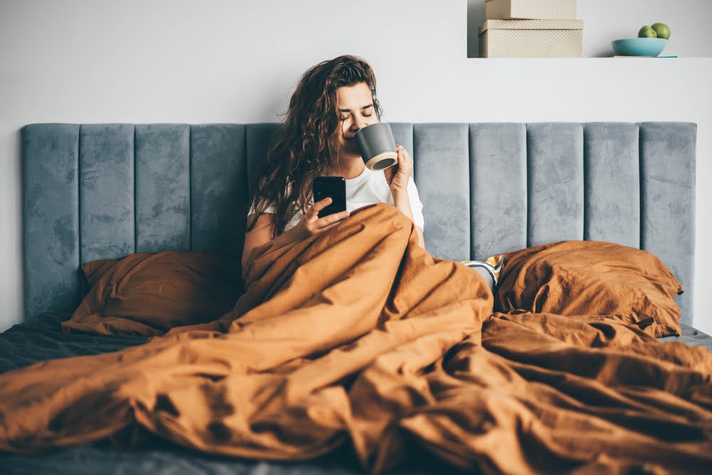 Woman drinking coffee in her bed and checking her smartphone. 
